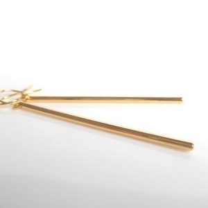 May include: A pair of gold-colored bar earrings. The earrings are simple, linear, and dangle from a hook. The bars are thin and have a polished finish, reflecting light. The background is white.