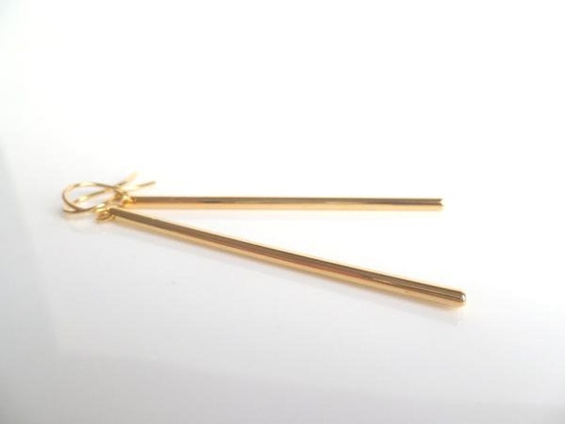 May include: A pair of gold-colored bar earrings. The earrings are simple, linear, and dangle from a hook. The bars are thin and have a polished finish, reflecting light. The background is white.