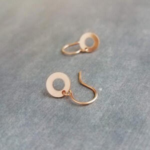 May include: Two rose gold earrings. Each earring features a small, circular disc with a hole in the center, connected to a curved ear wire. The earrings are displayed on a gray surface.