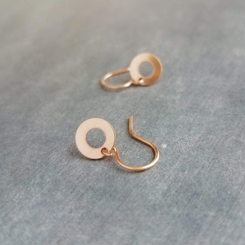 May include: Two rose gold earrings. Each earring features a small, circular disc with a hole in the center, connected to a curved ear wire. The earrings are displayed on a gray surface.