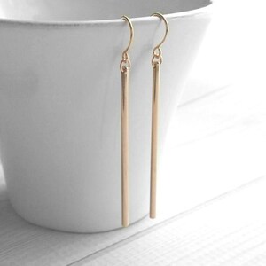 May include: A pair of gold-colored, linear drop earrings. Each earring features a simple, straight bar suspended from a curved ear wire. The earrings are displayed against a white background, highlighting their minimalist design and metallic sheen.