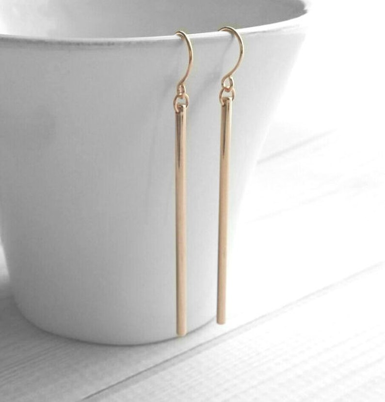May include: A pair of gold-colored, linear drop earrings. Each earring features a simple, straight bar suspended from a curved ear wire. The earrings are displayed against a white background, highlighting their minimalist design and metallic sheen.