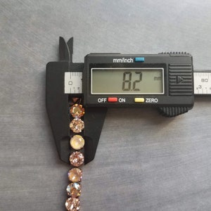 May include: A digital caliper measuring a strand of round, light brown gemstones. The digital display shows 8.2 mm. The gemstones are set in a row, with a metallic setting. The background is a gray surface.