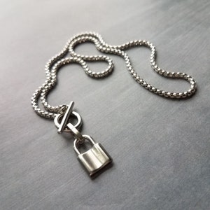 May include: A silver-colored necklace featuring a small padlock pendant. The necklace has a chain-link design and a toggle clasp. The padlock pendant is rectangular with a curved top. The necklace is displayed on a gray surface.