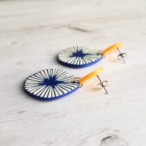 May include: A pair of handmade earrings featuring a navy blue oval with a white sunburst design. The earrings have orange accents and silver-colored posts. These unique earrings are a stylish accessory.