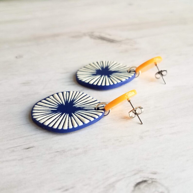 May include: A pair of handmade earrings featuring a navy blue oval with a white sunburst design. The earrings have orange accents and silver-colored posts. These unique earrings are a stylish accessory.