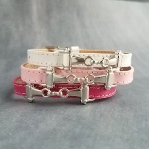 May include: Stack of three leather bracelets in white, light pink, and fuchsia. Each bracelet features a silver-colored metal bit design. The bracelets are stacked on top of each other, showcasing the different colors and designs.