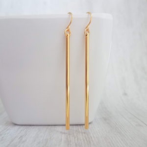 May include: A pair of gold-colored, long, straight bar earrings. The earrings hang from a simple hook. The earrings are displayed against a white background.