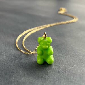 May include: A lime green gummy bear pendant necklace with a gold-colored chain. The gummy bear charm is glossy and detailed, with a small loop for the chain to pass through. The chain is delicate and appears to be made of gold.