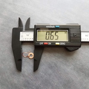May include: A copper-colored earring with a circular design, measuring 0.65 inches. The earring is displayed next to a digital caliper, which is measuring the earring's diameter. The caliper is black with a digital display.