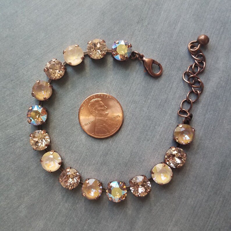 May include: A delicate bracelet featuring round, champagne-colored crystals with iridescent accents. The crystals are set in a copper-toned metal, connected by a thin wire. The bracelet includes a lobster clasp and an adjustable chain. A penny is next to the bracelet.