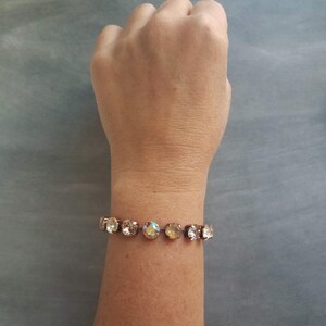 May include: A gold-toned bracelet featuring a series of round, iridescent crystals. The crystals are set closely together, creating a continuous band of sparkle. The bracelet is shown on a wrist against a gray background.