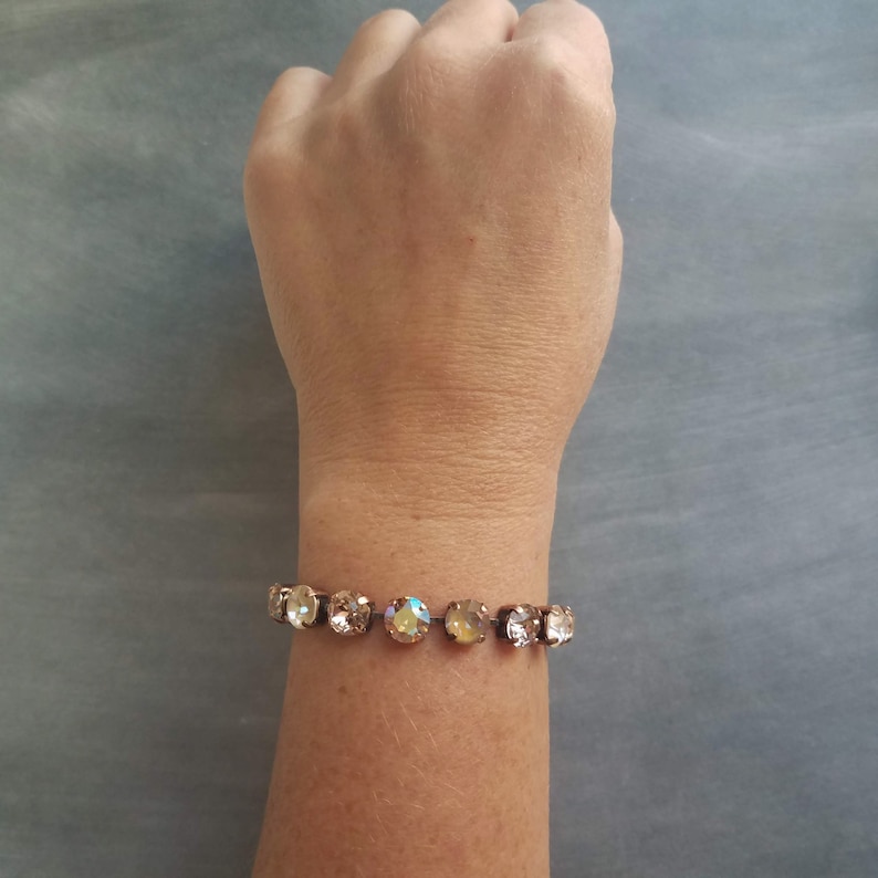 May include: A gold-toned bracelet featuring a series of round, iridescent crystals. The crystals are set closely together, creating a continuous band of sparkle. The bracelet is shown on a wrist against a gray background.