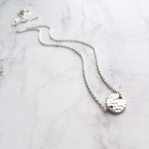 May include: A silver necklace featuring a hammered-texture pendant. The pendant is a small, circular disc attached to a fine chain. The necklace is shown on a white and grey marble surface.
