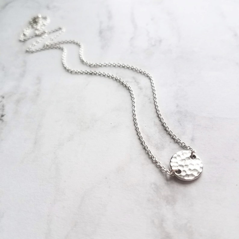 May include: A silver necklace featuring a hammered-texture pendant. The pendant is a small, circular disc attached to a fine chain. The necklace is shown on a white and grey marble surface.