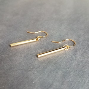 Peut inclure: Une paire de boucles d'oreilles en forme de barre dor&eacute;e. Chaque boucle d'oreille est compos&eacute;e d'une barre droite et rectangulaire suspendue &agrave; un crochet incurv&eacute;. Les boucles d'oreilles sont pr&eacute;sent&eacute;es sur un fond gris, soulignant leur design minimaliste.