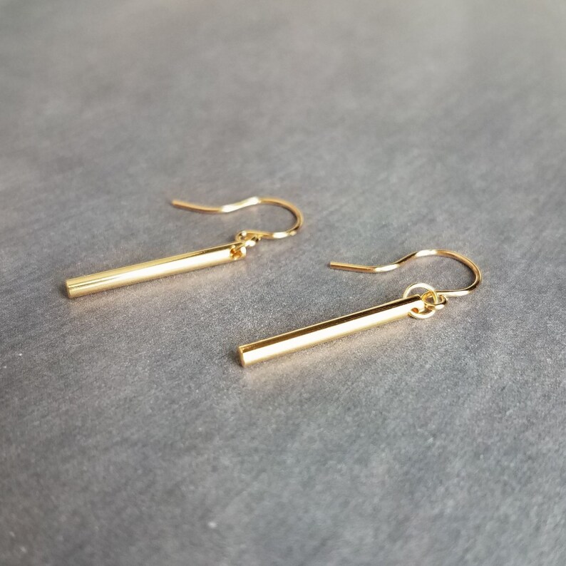 Peut inclure: Une paire de boucles d'oreilles en forme de barre dor&eacute;e. Chaque boucle d'oreille est compos&eacute;e d'une barre droite et rectangulaire suspendue &agrave; un crochet incurv&eacute;. Les boucles d'oreilles sont pr&eacute;sent&eacute;es sur un fond gris, soulignant leur design minimaliste.