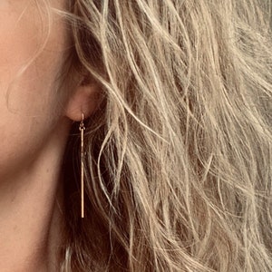 May include: Gold-colored, linear drop earrings. The earrings are long, thin, and dangle from a small hook. The earrings are shown against a background of wavy, light-colored hair.