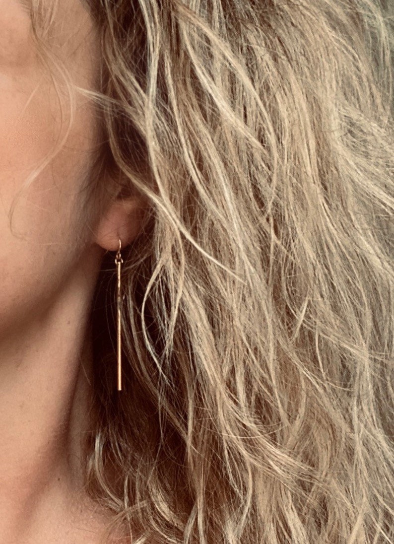 May include: Gold-colored, linear drop earrings. The earrings are long, thin, and dangle from a small hook. The earrings are shown against a background of wavy, light-colored hair.