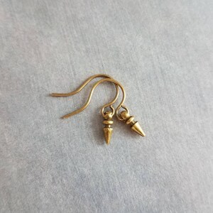 May include: A pair of small, antique gold-colored earrings. Each earring features a simple hook and a small, cone-shaped pendant. The earrings are displayed against a gray background.