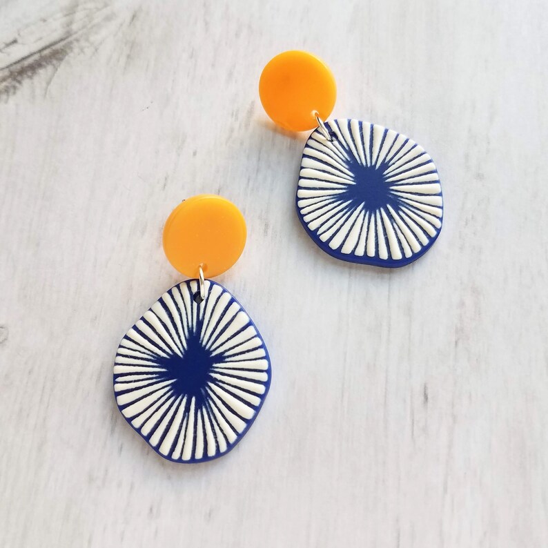 May include: A pair of earrings featuring a bright yellow circular stud and a navy blue, abstract, teardrop-shaped pendant. The pendant has a white, radial design. The earrings are displayed on a light wood-grain surface.
