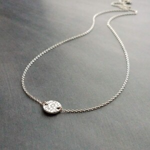 May include: A delicate silver necklace with a hammered, circular pendant. The pendant is attached to a fine chain, creating a minimalist design. The necklace is displayed on a dark grey surface.
