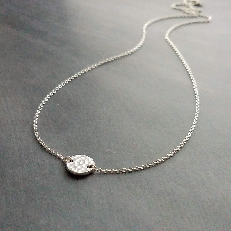 May include: A delicate silver necklace with a hammered, circular pendant. The pendant is attached to a fine chain, creating a minimalist design. The necklace is displayed on a dark grey surface.