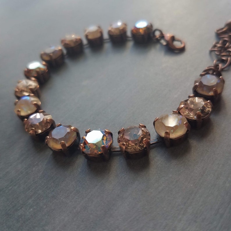 May include: A close-up of a bracelet featuring iridescent, round gemstones set in a bronze-colored metal. The gemstones have a range of colors, including gold, peach, and hints of blue. The bracelet is on a dark gray surface.