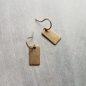 May include: A pair of rectangular brass earrings with curved ear wires. The earrings have a simple, minimalist design and a warm, metallic finish. The earrings are displayed on a gray surface.
