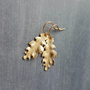 May include: A pair of gold-colored leaf earrings. The earrings are shaped like oak leaves with detailed veining. The leaves hang from simple gold-colored ear wires. The earrings are displayed against a gray background.
