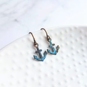 Könnte beinhalten: Ein Paar kleiner, ankerförmiger Ohrringe mit einem verwitterten, türkisfarbenen und bronzefarbenen Finish. Die Ohrringe hängen an bronzefarbenen Ohrhaken. Die Ohrringe werden auf einer weißen, strukturierten Oberfläche präsentiert.