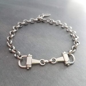 May include: Silver-toned bracelet featuring a horse bit design. The bracelet has a chain-link style with a central horse bit clasp. The metal has a polished finish, reflecting light. A simple, elegant piece of jewelry.