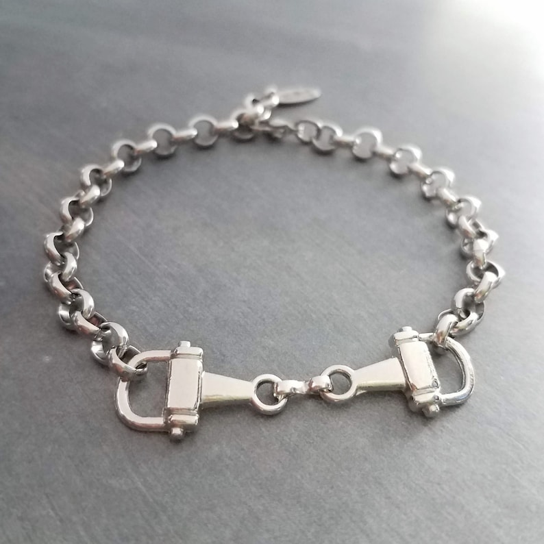 May include: Silver-toned bracelet featuring a horse bit design. The bracelet has a chain-link style with a central horse bit clasp. The metal has a polished finish, reflecting light. A simple, elegant piece of jewelry.