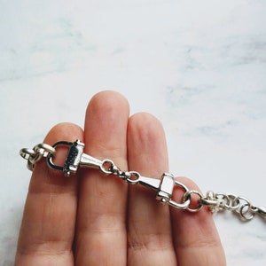 May include: Silver-toned equestrian-style bracelet featuring a horse bit design. The bracelet has a series of rounded links and a central bit element with a hinged design. The metal has a polished finish, reflecting light.