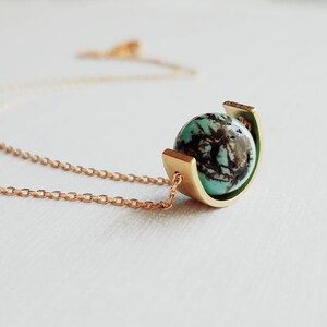 May include: A necklace featuring a round turquoise bead with dark brown matrix patterns, held within a curved gold-toned metal cradle. The pendant is attached to a delicate gold-toned chain that extends outward from the sides of the metal setting.
