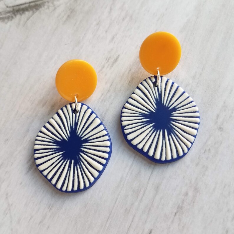 May include: A pair of geometric earrings. Each earring features a small, round, yellow stud connected to a navy blue, diamond-shaped pendant with white radial lines. The earrings are designed with a modern, abstract aesthetic.