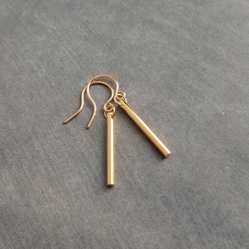 Peut inclure: Une paire de boucles d'oreilles en forme de barres dor&eacute;es. Chaque boucle d'oreille est compos&eacute;e d'une barre droite suspendue &agrave; un crochet incurv&eacute;. Les boucles d'oreilles sont pr&eacute;sent&eacute;es sur un fond gris, soulignant leur design minimaliste et leur &eacute;clat m&eacute;tallique.