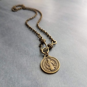 May include: An antique bronze-toned necklace featuring a round pendant with an image of a saint and text. The pendant hangs from a delicate chain with a decorative clasp. The necklace is displayed on a gray surface.