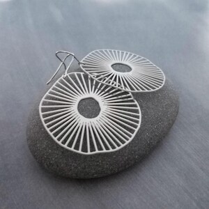 Könnte beinhalten: Ein Paar silberfarbene, offene Ohrringe mit radialem Design, die Sonnenstrahlen ähneln. Die Ohrringe werden auf einem grauen Stein vor grauem Hintergrund präsentiert. Die Ohrringe haben eine kreisförmige Form mit einem zentralen Loch und radialen Linien.