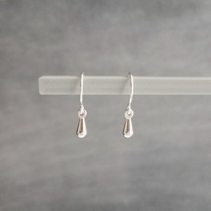May include: A pair of small, silver teardrop earrings hanging from a white bar against a gray background. The earrings have a simple, elegant design with a smooth, polished finish. The hooks are thin and curved.