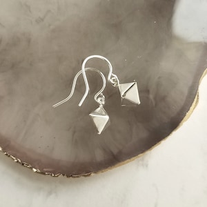 May include: A pair of silver geometric earrings. Each earring features a small, three-dimensional diamond shape suspended from a curved ear wire. The earrings are displayed on a light gray and brown surface.