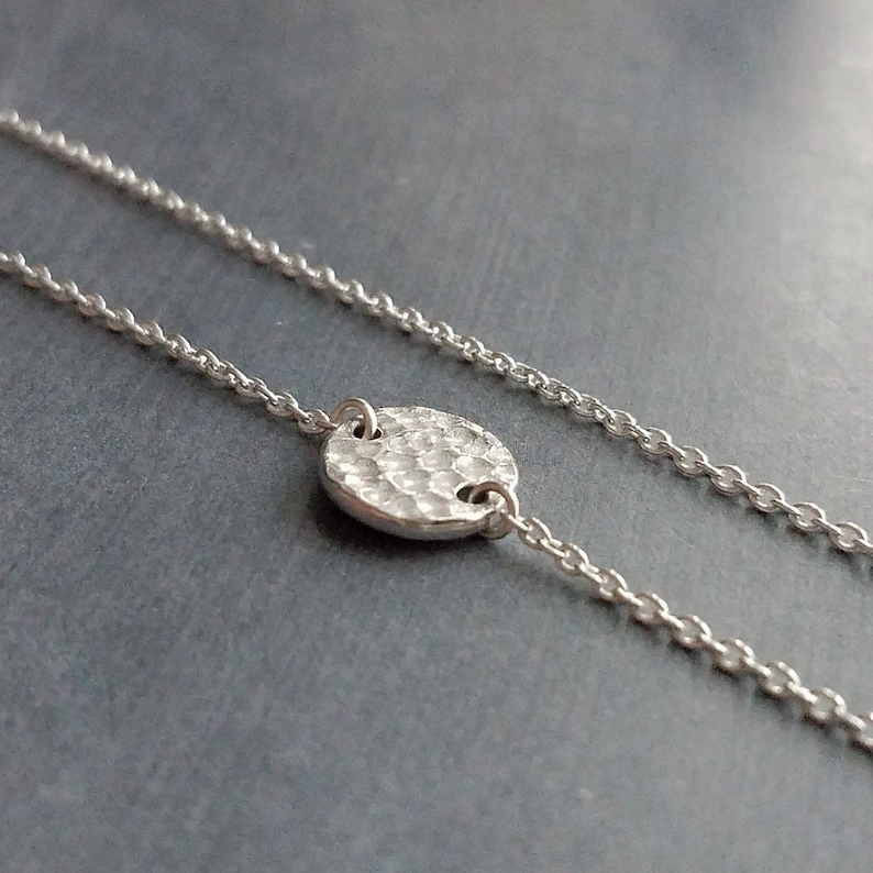 May include: A silver necklace featuring a hammered disc pendant. The pendant is round with a textured surface, attached to a fine chain. The necklace is set against a dark grey backdrop.