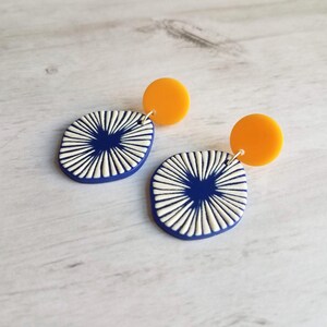 May include: A pair of modern earrings. Each earring features a navy blue, oval-shaped pendant with a white, sunburst design, connected to a small, round, orange stud. The earrings are displayed on a light-colored, wood-grain surface.