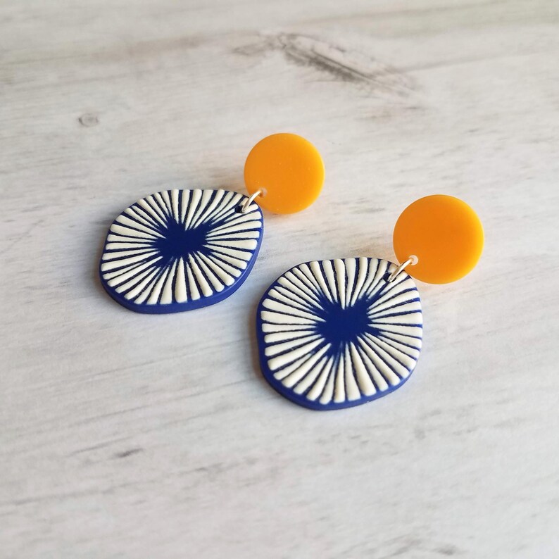 May include: A pair of modern earrings. Each earring features a navy blue, oval-shaped pendant with a white, sunburst design, connected to a small, round, orange stud. The earrings are displayed on a light-colored, wood-grain surface.