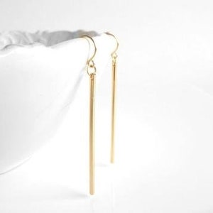 May include: A pair of gold-colored, linear drop earrings. The earrings are long, slender, and hang vertically. The earrings are suspended from a curved hook. The background is white, and a white ceramic bowl is partially visible.