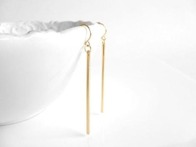 May include: A pair of gold-colored, linear drop earrings. The earrings are long, slender, and hang vertically. The earrings are suspended from a curved hook. The background is white, and a white ceramic bowl is partially visible.