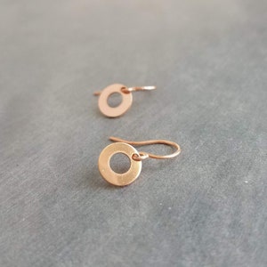 May include: A pair of small, copper-colored circle earrings. Each earring features a circular disc with a hole in the center, suspended from a simple hook. The earrings are displayed on a gray surface, showcasing their minimalist design and warm metallic hue.