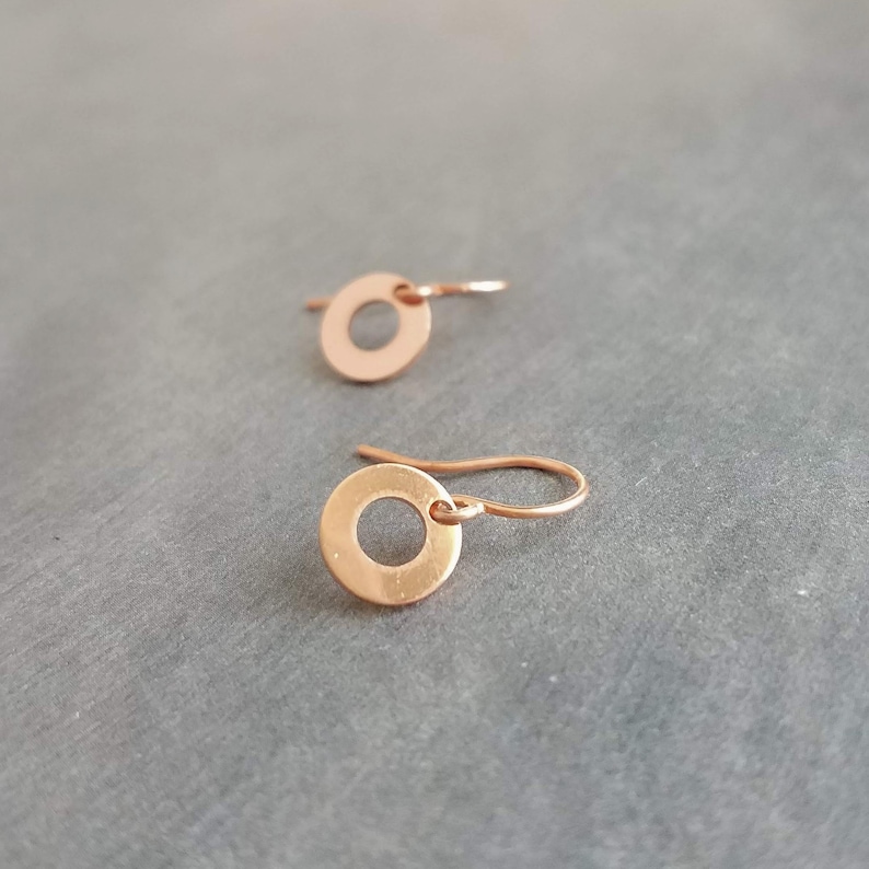 May include: A pair of small, copper-colored circle earrings. Each earring features a circular disc with a hole in the center, suspended from a simple hook. The earrings are displayed on a gray surface, showcasing their minimalist design and warm metallic hue.