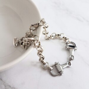 May include: A silver-toned bracelet featuring a series of connected circular links and two decorative horse bit charms. The bracelet is displayed on a white surface, with a portion of a white bowl visible in the background.