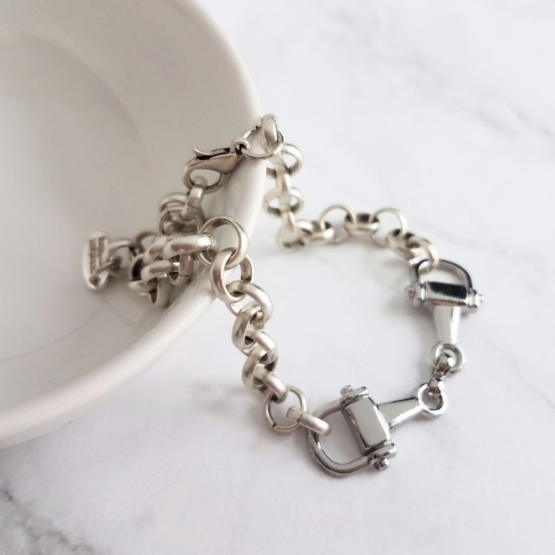 May include: A silver-toned bracelet featuring a series of connected circular links and two decorative horse bit charms. The bracelet is displayed on a white surface, with a portion of a white bowl visible in the background.
